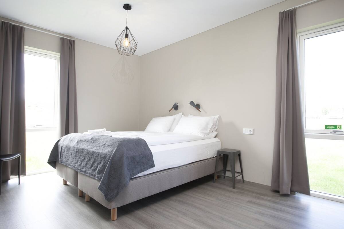 Une chambre moderne avec un lit, une table de chevet, un luminaire suspendu et de grandes fenêtres avec des rideaux.