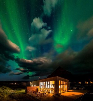Un edificio con zona de bar y grandes ventanales, bajo un cielo nocturno con auroras boreales visibles.