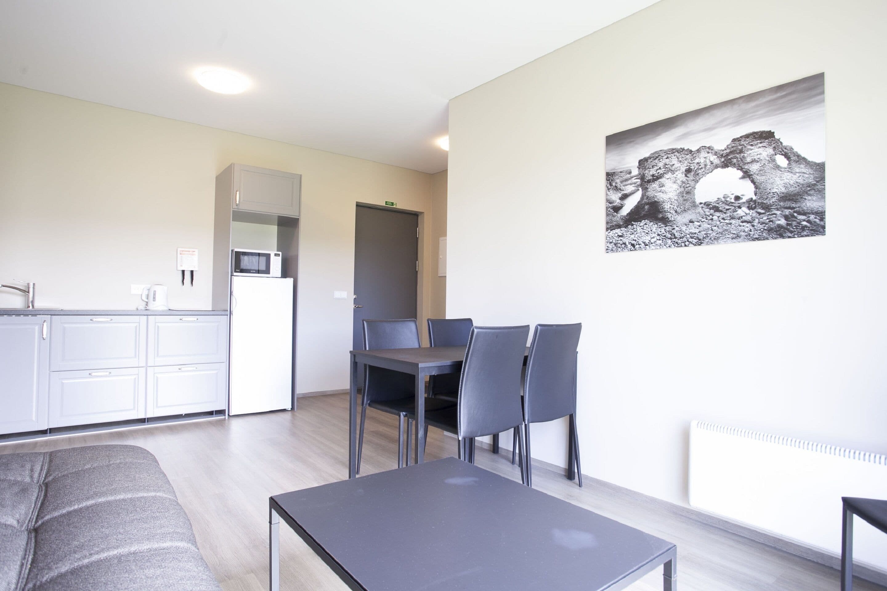 Una cocina moderna con armarios blancos, un microondas y un refrigerador. Hay una zona de comedor con una mesa y sillas. Un sofá gris se encuentra en primer plano.