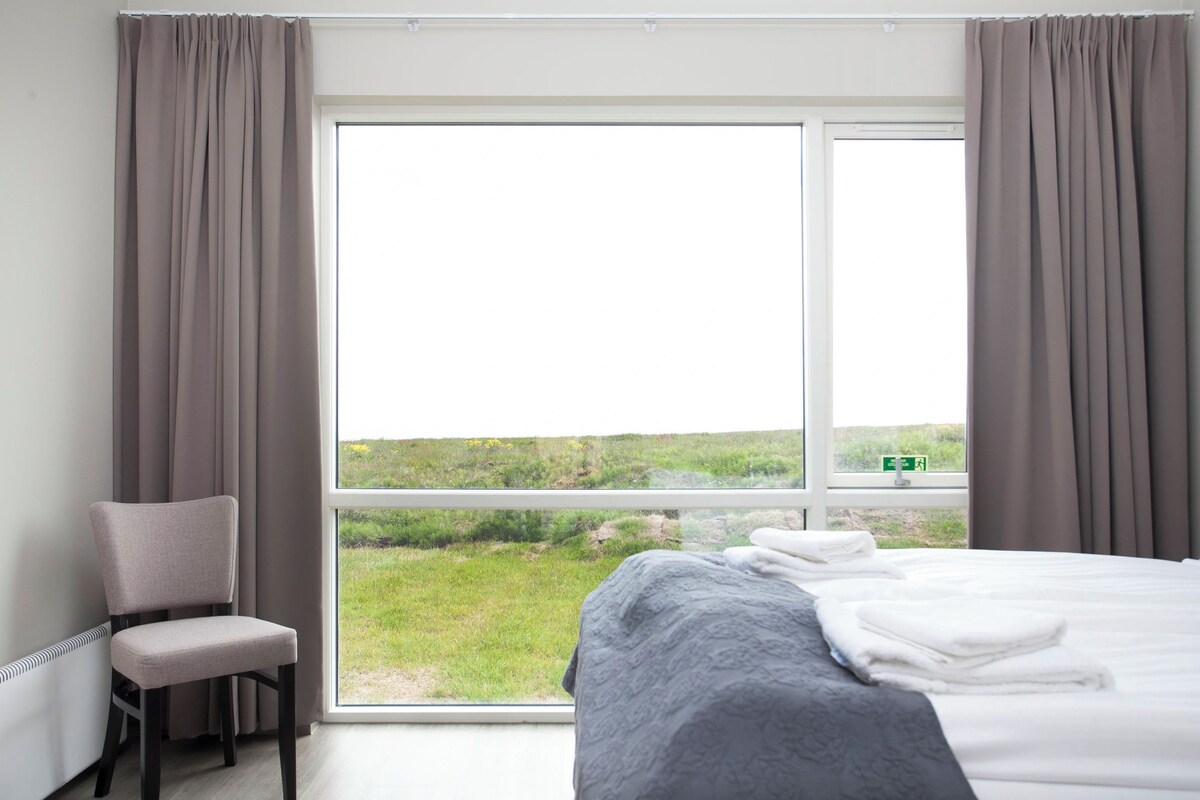 A hotel room with a large window, a bed, a chair, and a view of a grassy landscape.