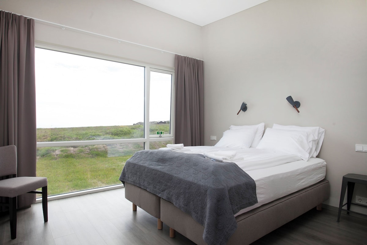 Un dormitorio moderno con una cama grande, una silla y vistas a un campo cubierto de hierba a través de un ventanal amplio.