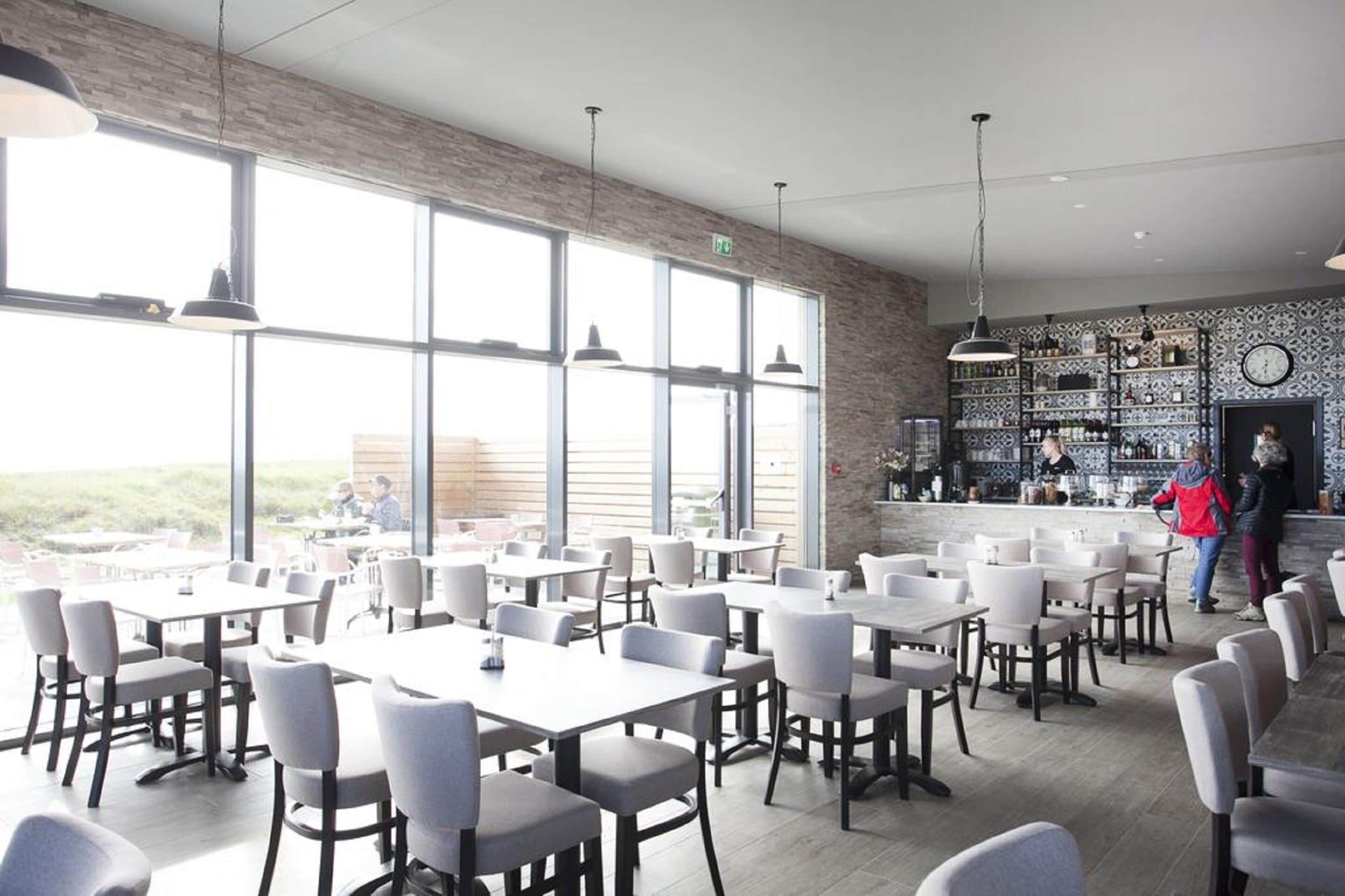 A spacious dining area with large windows, modern furniture, and a bar area visible in the background.