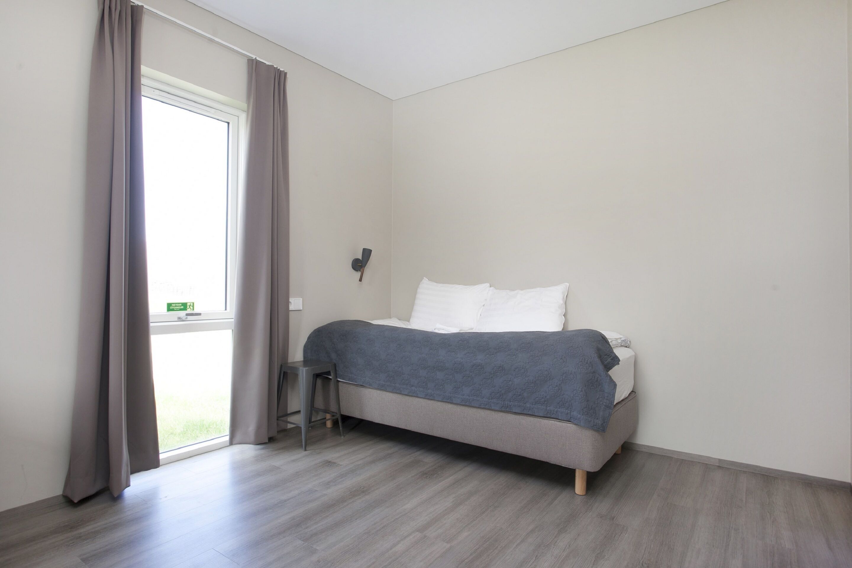 A single bed with a grey blanket, a small stool, and a window with curtains.