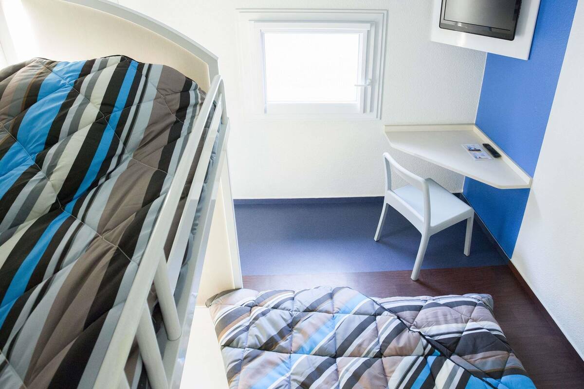 Una habitación con cama, escritorio, silla, televisor montado en la pared y ventana.