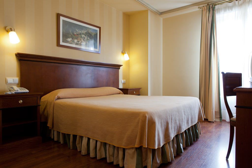 Habitación de hotel con una cama grande, cabecera de madera, dos mesitas de noche, un buró y una silla.