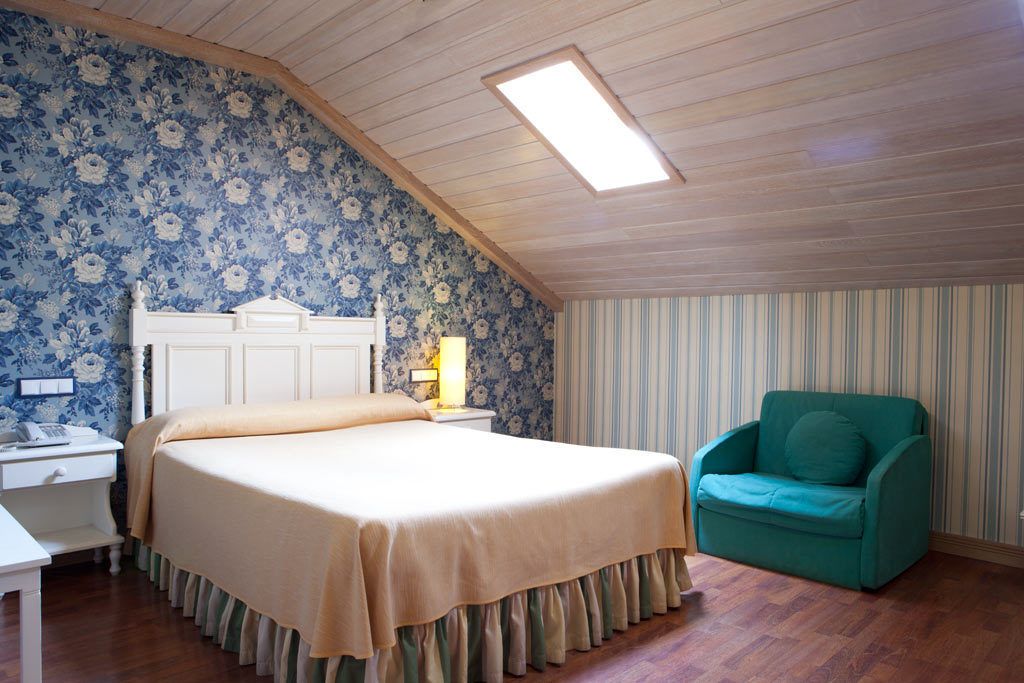 A bedroom with a bed, a nightstand, a lamp, a chair, and a window with a skylight.