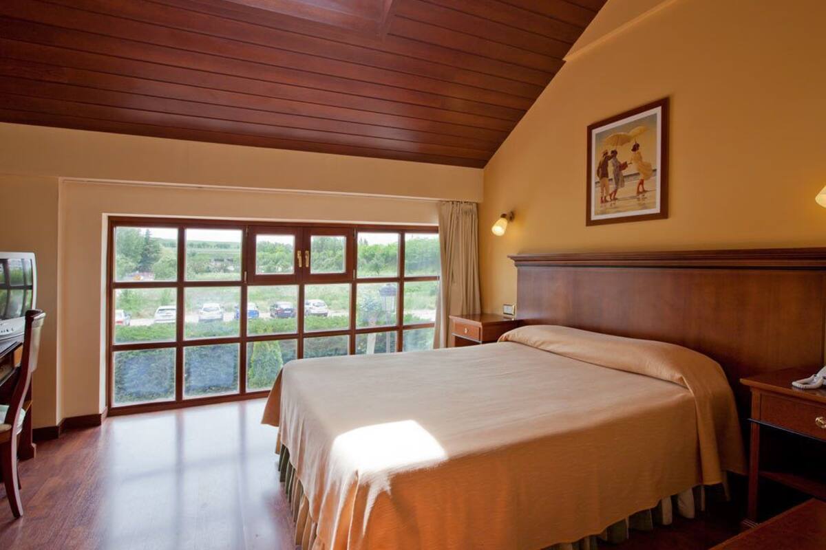 Une chambre d’hôtel avec un grand lit, un bureau avec une chaise, un piano et une vue sur un parking, visible à travers une grande fenêtre.