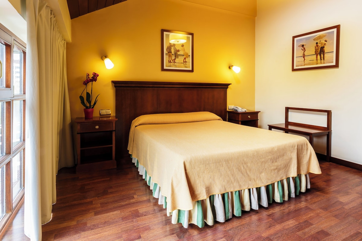 A neatly arranged hotel room with a large bed, a bedside table, a nightstand, a lamp, and two framed pictures on the wall.