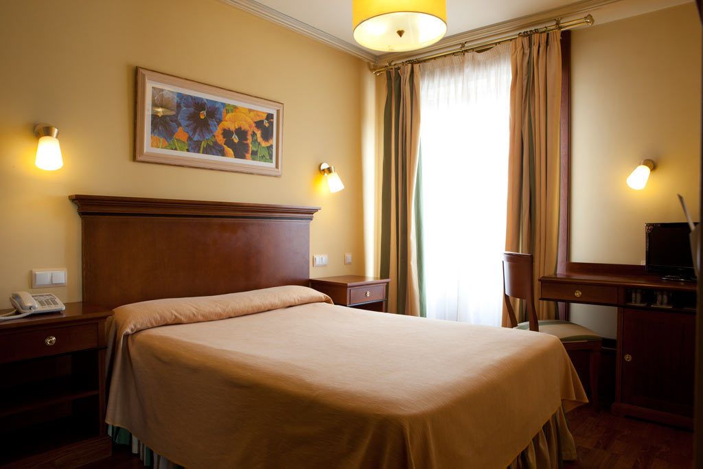 A hotel room with a large bed, a desk with a chair, a telephone, and a painting on the wall.