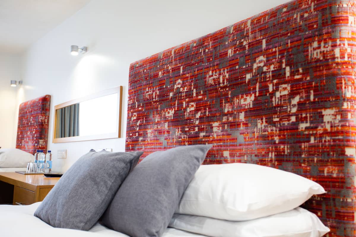 A hotel room with a bed, pillows, a nightstand, a mirror, and a patterned headboard.