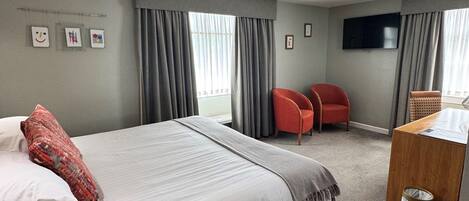 Una habitación de hotel con una cama, dos sillas rojas, un escritorio y un televisor en la pared.