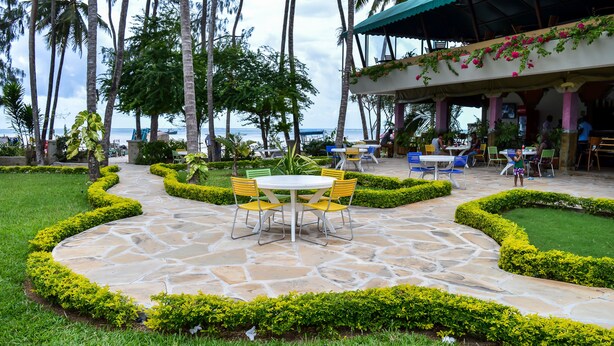 A patio area with stone flooring, yellow chairs, and tables, surrounded by well-manicured hedges and palm trees. - Lido Beach Resort (Mombasa)