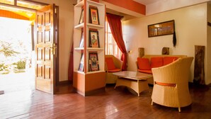 A room with a wooden floor, a sofa with orange cushions, a wicker chair, a bookshelf with framed pictures, and a door leading outside. - Magharibi Garden Hotel (Kisii)