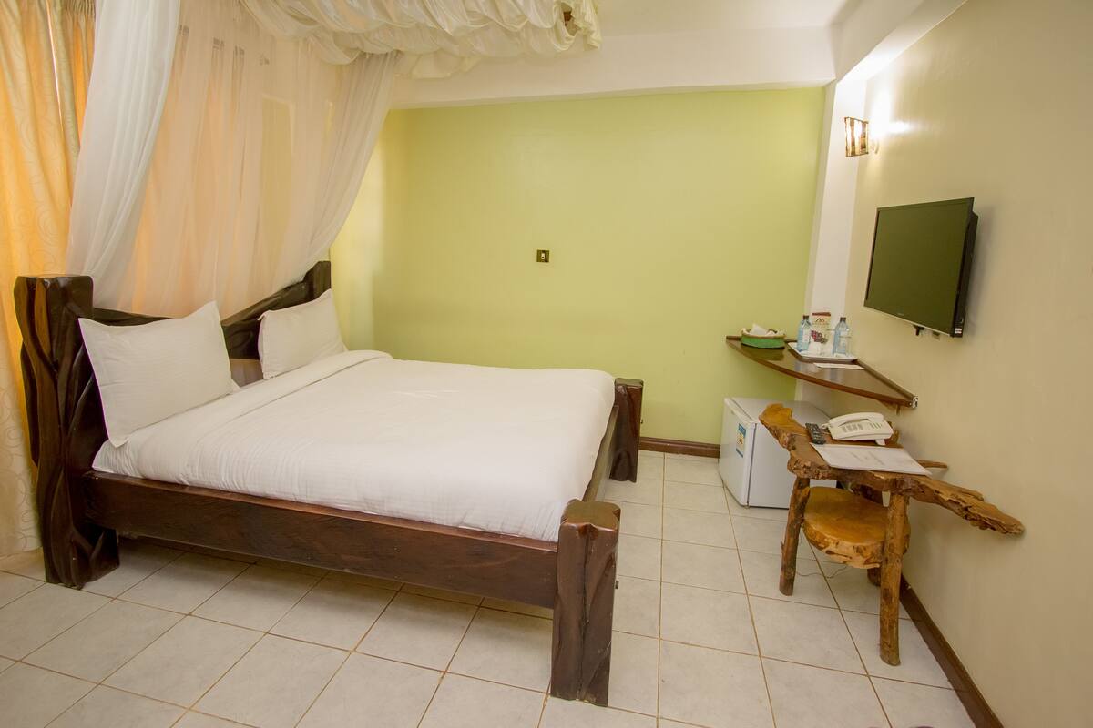 Un dormitorio con cama de madera, una mesita de noche con teléfono, televisor montado en la pared y un escritorio pequeño con silla.