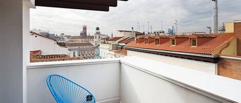 A balcony with two blue chairs and a small wooden table, overlooking a cityscape with red-tiled roofs and historic buildings.
