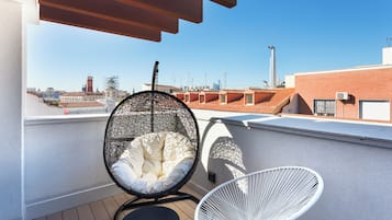 A rooftop terrace with a hanging chair, a white chair, and a view of the cityscape.