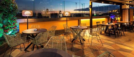 An outdoor dining area with wicker chairs and tables, a glass wall with a city view, and a sign reading "Nulsinob".