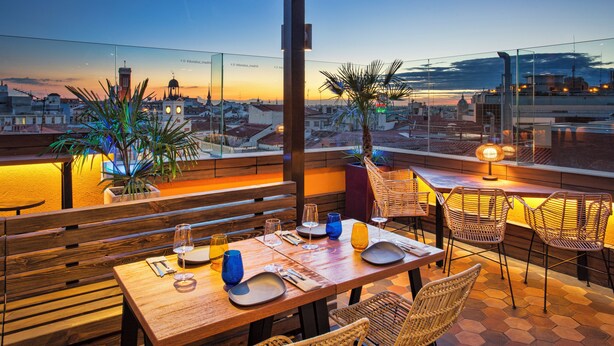 Eine Dachterrasse mit einem Holztisch, gedecktem Esstisch, Blumentöpfen und Blick auf die Stadtlandschaft im Abenddämmerung.