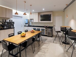 Een moderne keuken met een houten tafel, stoelen en een schaal met fruit. Er is een magnetron, koffieapparaat en een televisie aan de muur bevestigd.