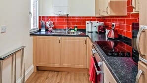 A compact kitchen with wooden cabinets, a black countertop, a red-tiled backsplash, a wall clock, and a window. - Conway Cottages (York)