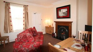 A cozy living room with a red floral sofa, a fireplace, a dining table set for two, and a framed picture on the wall. - Conway Cottages (York)