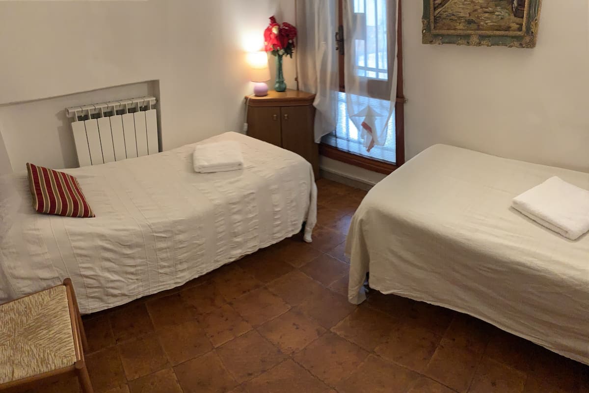 Quarto com duas camas de solteiro, uma cadeira, um radiador, uma mesa de cabeceira com luminária e flores, e uma janela com vista.