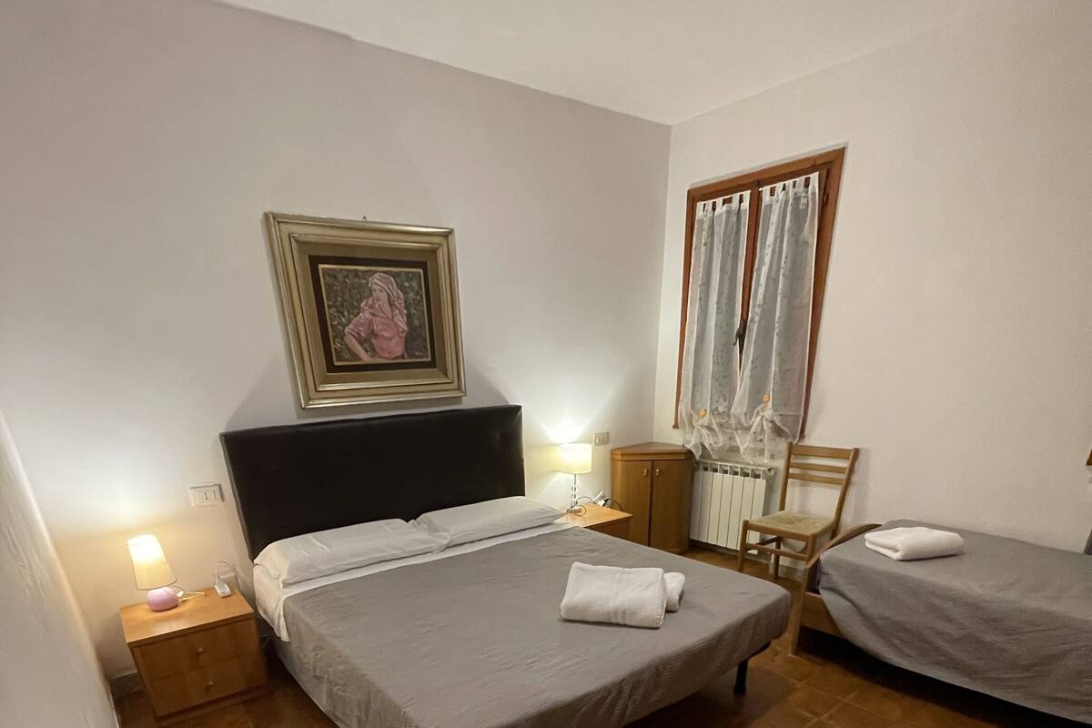 Um quarto com cama, mesa de cabeceira, cadeira, janela com cortinas e um quadro na parede.