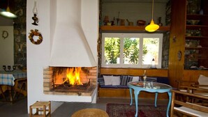 A cozy living room with a fireplace, a blue table, and a view of greenery through a window. - Villa Aggeliki (Eretria)