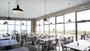 A spacious dining area with multiple tables and chairs, large windows offering a view of the beach and cars outside, and hanging pendant lights. - Arnarstapi Cottages (Arnarstapi)