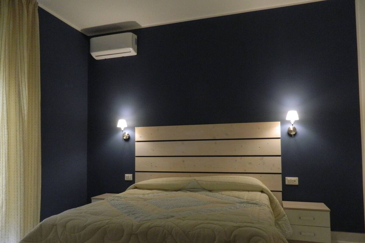 A bedroom with a wooden headboard, two wall-mounted lights, a bed with a quilted bedspread, a bedside table, and an air conditioning unit.
