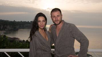A couple standing on a balcony overlooking a coastal landscape with cliffs and a body of water.