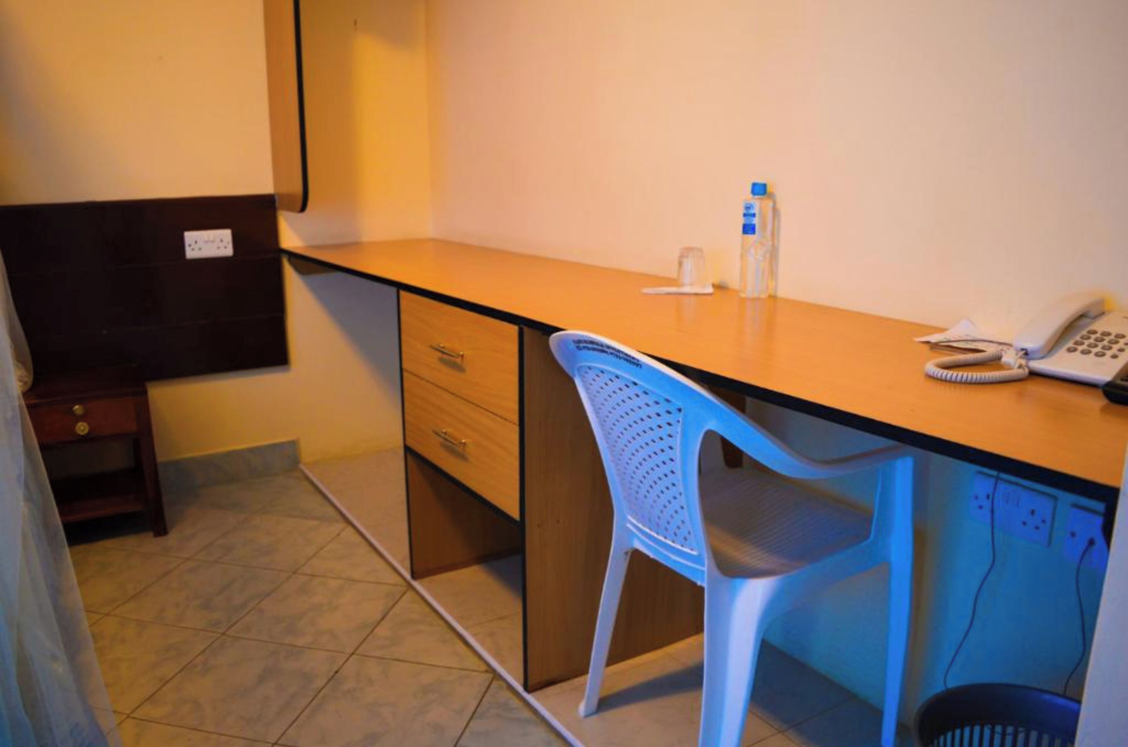 Habitación de hotel con un escritorio de madera, una silla de plástico azul, un teléfono y un pequeño cubo de basura.