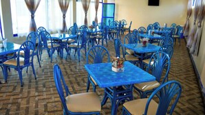 Un comedor con mesas y sillas azules, piso estampado y ventanas grandes con cortinas.