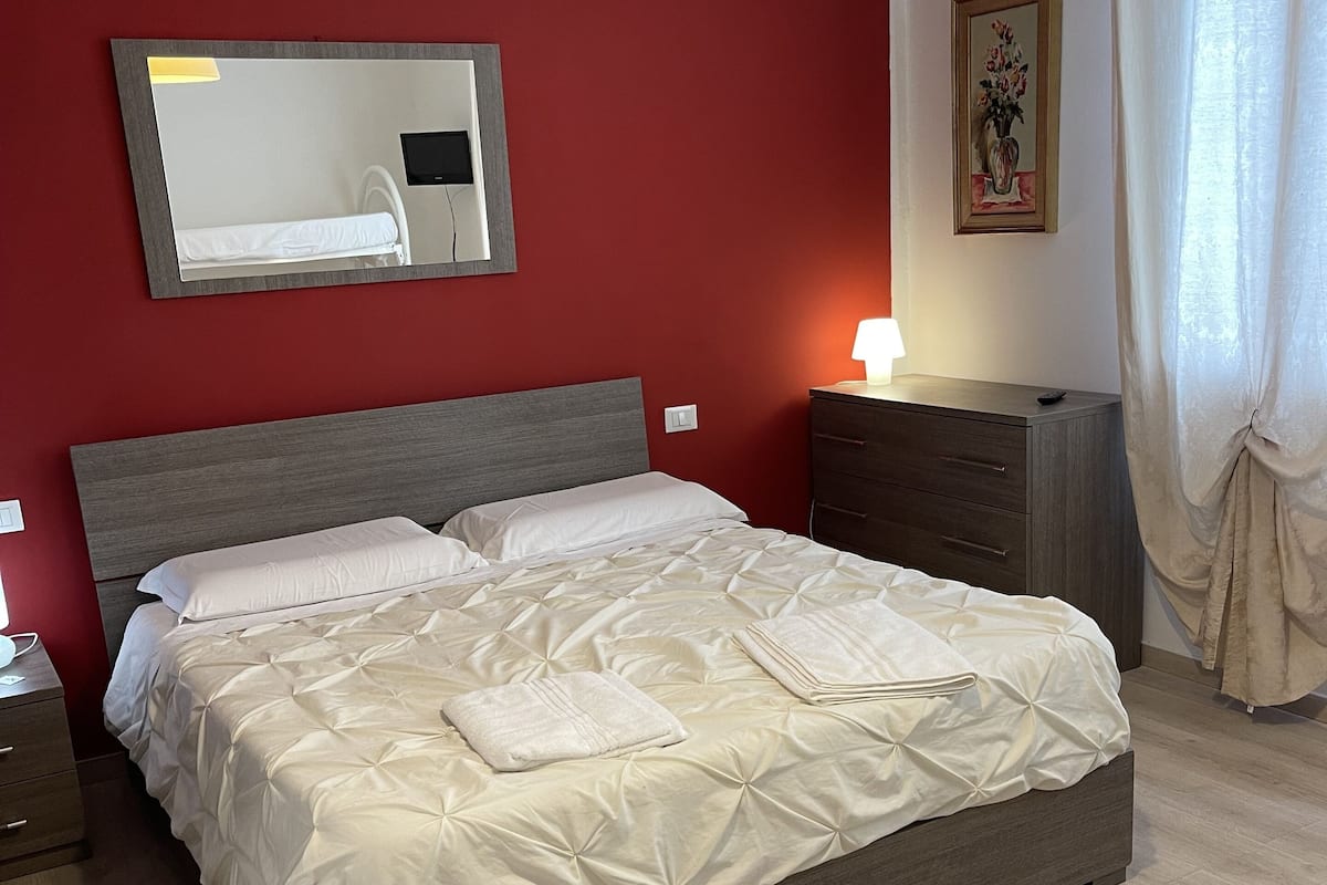 A neatly made bed with a grey headboard, a bedside table with a lamp, and a wooden dresser.