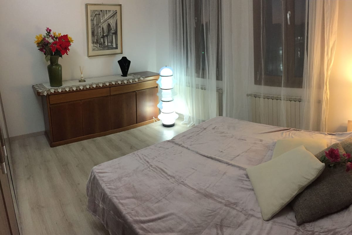A bedroom with a bed, bedside table, lamp, and a vase with flowers.