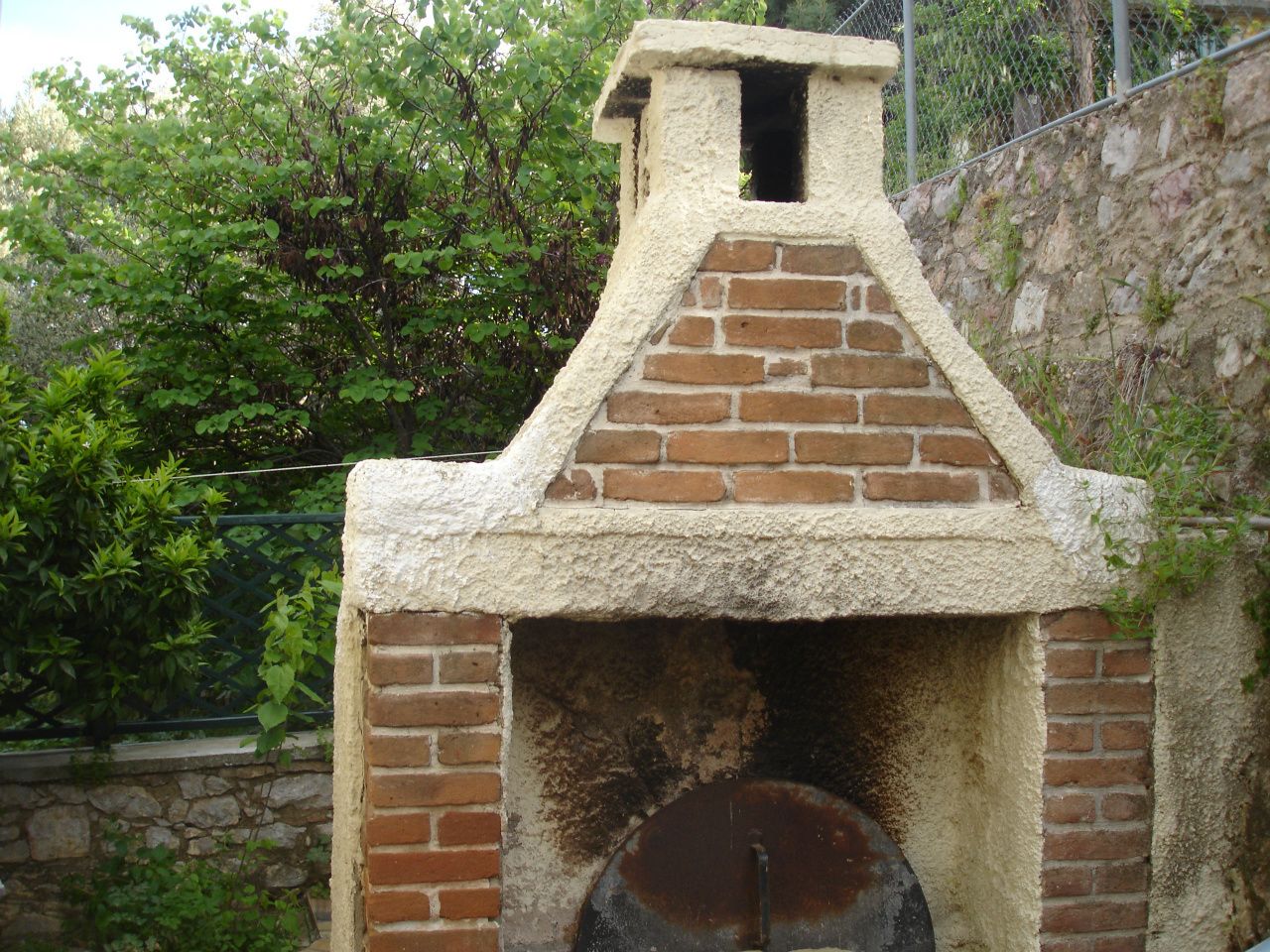 A brick oven with a chimney, surrounded by greenery and a stone wall.