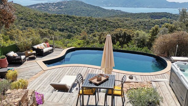 Una piscina con jacuzzi, rodeada de una cubierta de madera, mobiliario exterior y una vista panorámica de colinas y un lago.