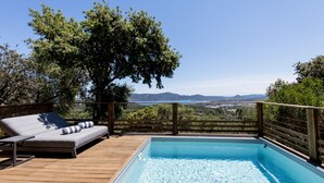 Una terraza de madera con piscina, un área de descanso con sofá y cojines, y vistas del paisaje.