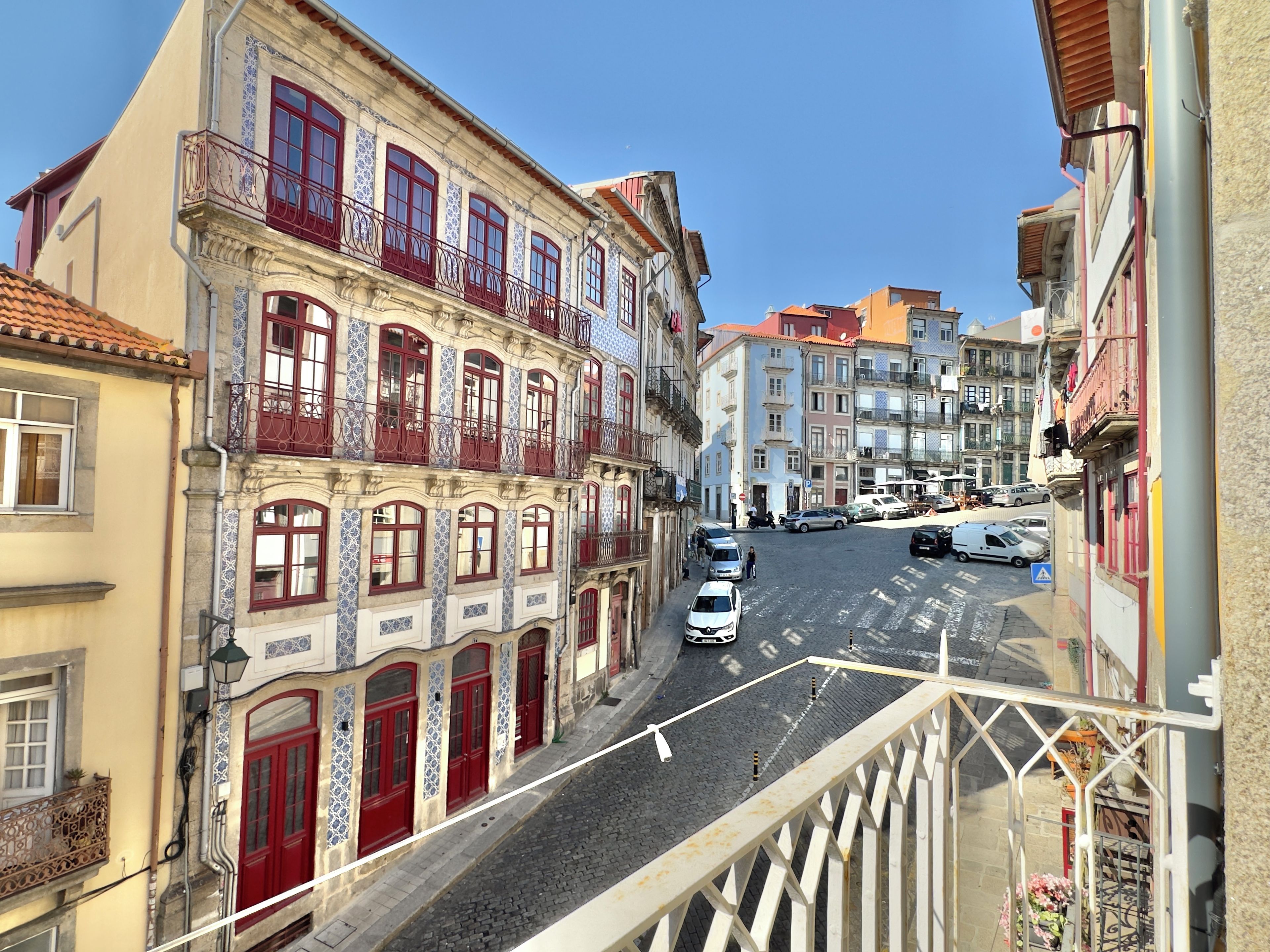 A street view with buildings featuring ornate facades and balconies, a cobblestone street, and parked cars.