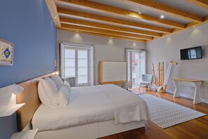 A modern bedroom with a large bed, wooden beams, a TV, and a desk. - YOUROPO - ExtendALL (Porto)