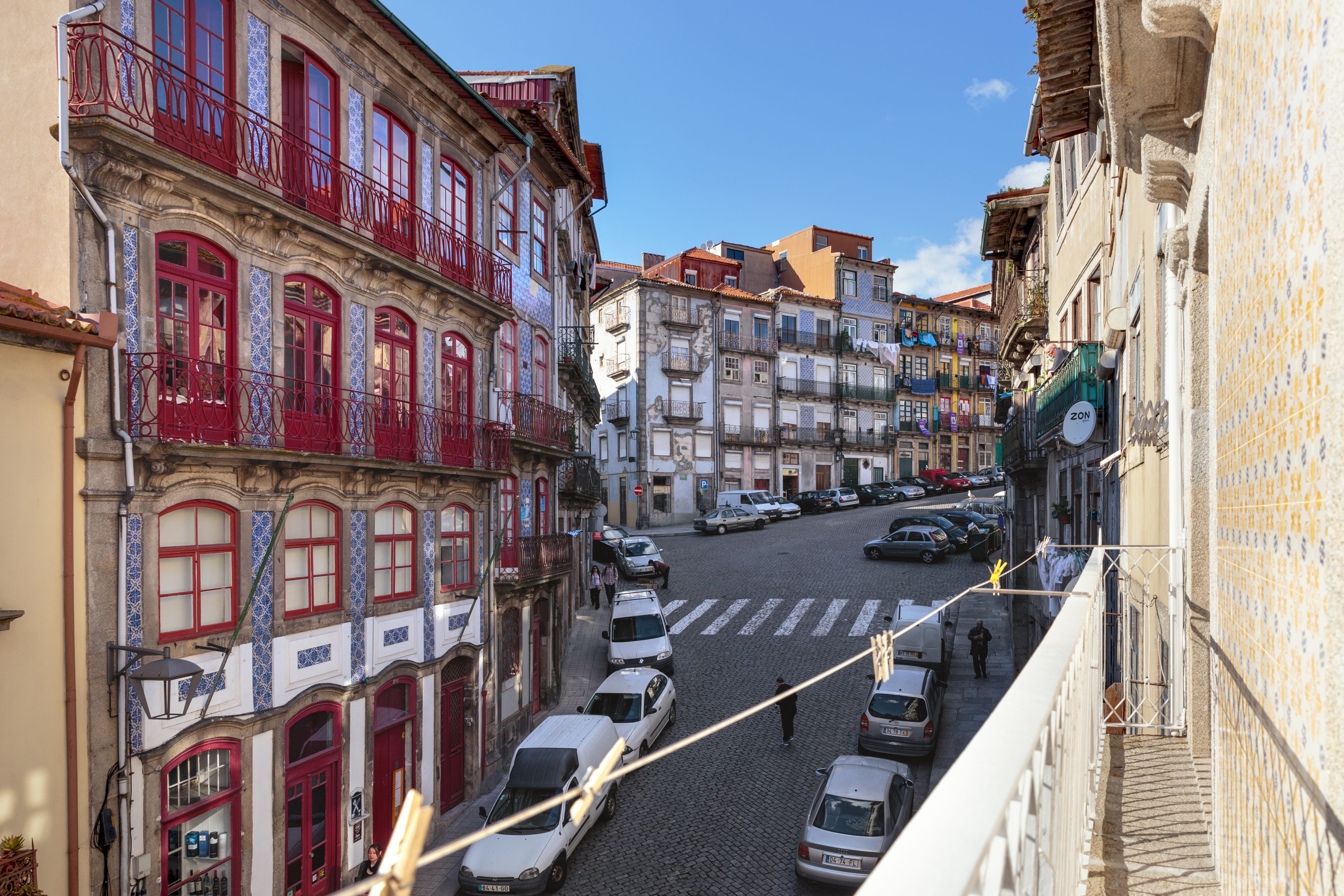 A narrow street with parked cars and buildings with red balconies and tiled facades.