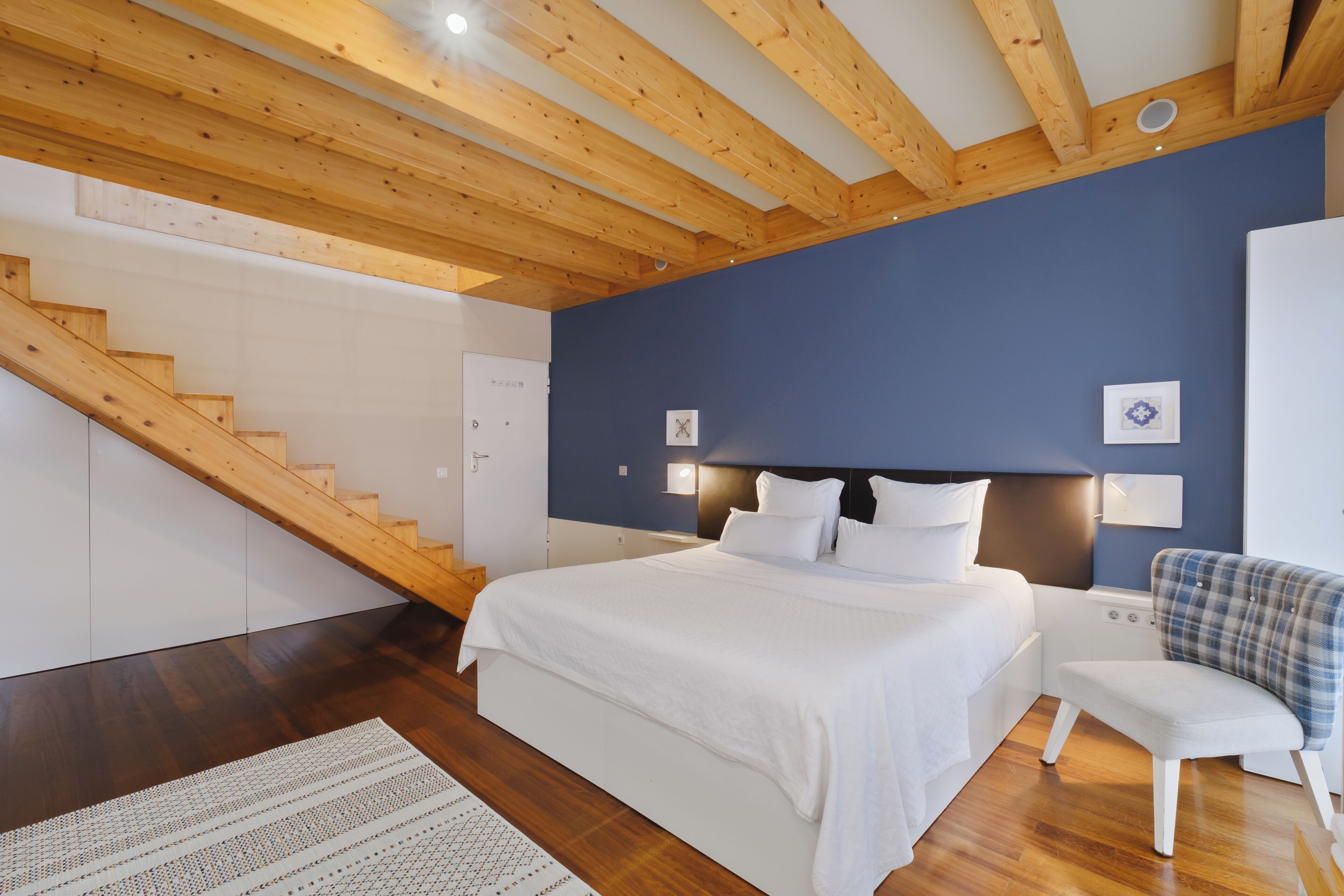 A bedroom with a wooden staircase, a bed with white bedding, a nightstand, and a chair.