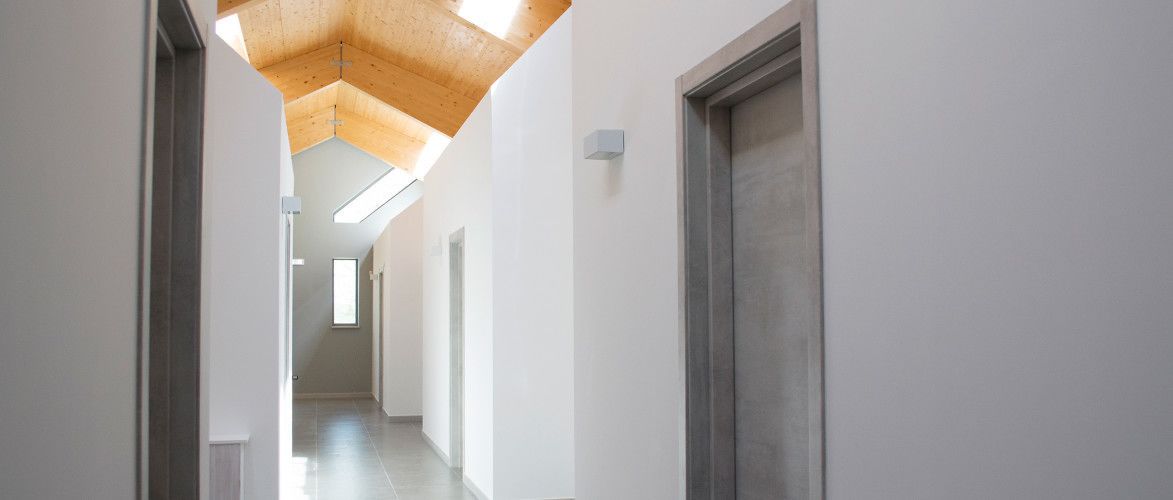 A long, narrow hallway with white walls and a wooden ceiling. There are doors on both sides and a window at the end.
