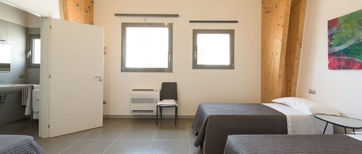 Habitación de hotel con dos camas, un escritorio y una silla. Dos ventanas dejan entrar luz natural.