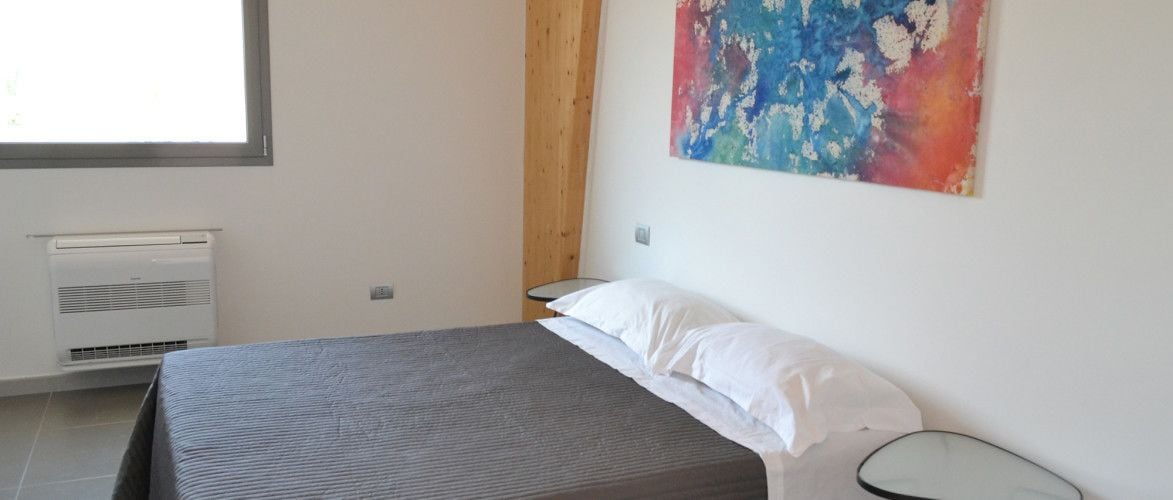 A bedroom with a bed, a nightstand, a wall-mounted air conditioner, and a colorful abstract painting.