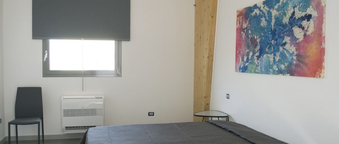 Un dormitorio con cama, una silla, aire acondicionado, ventana y un cuadro en la pared.