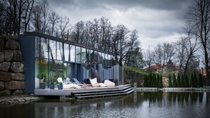 Una moderna casa flotante con fachada de vidrio, mobiliario exterior y un estanque al fondo.