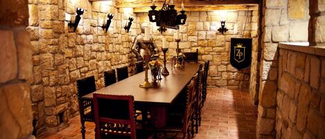 A stone-walled dining room with a long wooden table, chairs, and a chandelier.