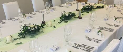 Una larga mesa de comedor dispuesta para un evento formal, con manteles blancos, cubiertos y copas.