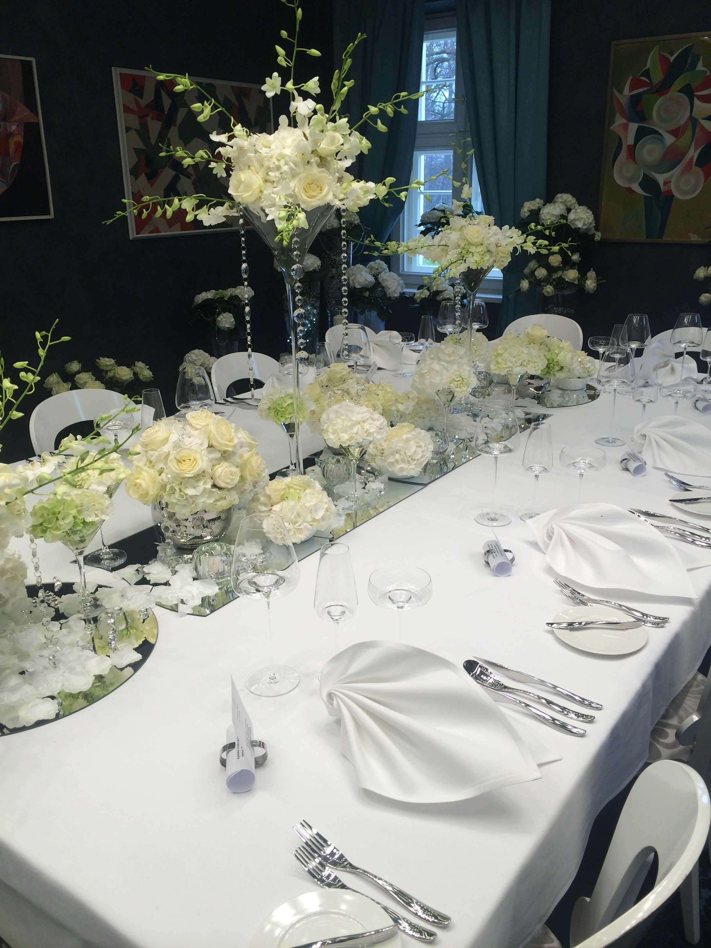 A long banquet table set with white tablecloths, silverware, and glassware, adorned with white flowers and greenery.